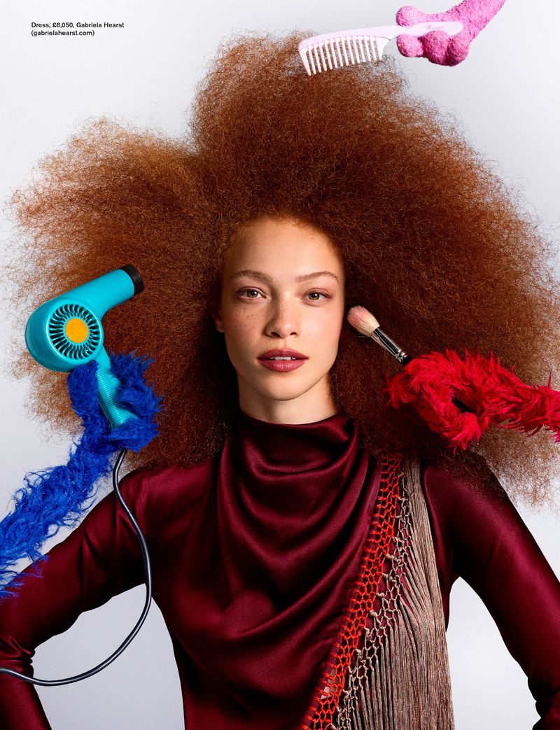 Tianna St. Louis with large auburn afro hair styled by fuzzy hands holding a hairdryer, brush, and comb against a white background.