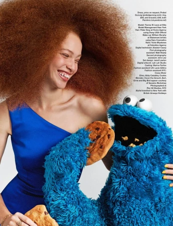 Model Tianna St. Louis in a blue one-shoulder dress smiling alongside a Cookie Monster puppet holding cookies against a white backdrop.