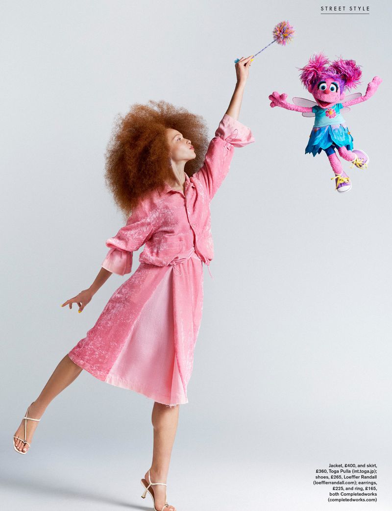 Model Tianna St. Louis wearing a pink dress and holding a wand toward a floating Abby Cadabby puppet against a white background.
