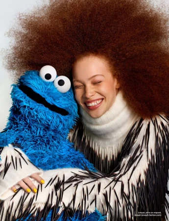 Model Tianna St. Louis smiling with voluminous auburn afro hair while embracing the Cookie Monster puppet on a white backdrop.