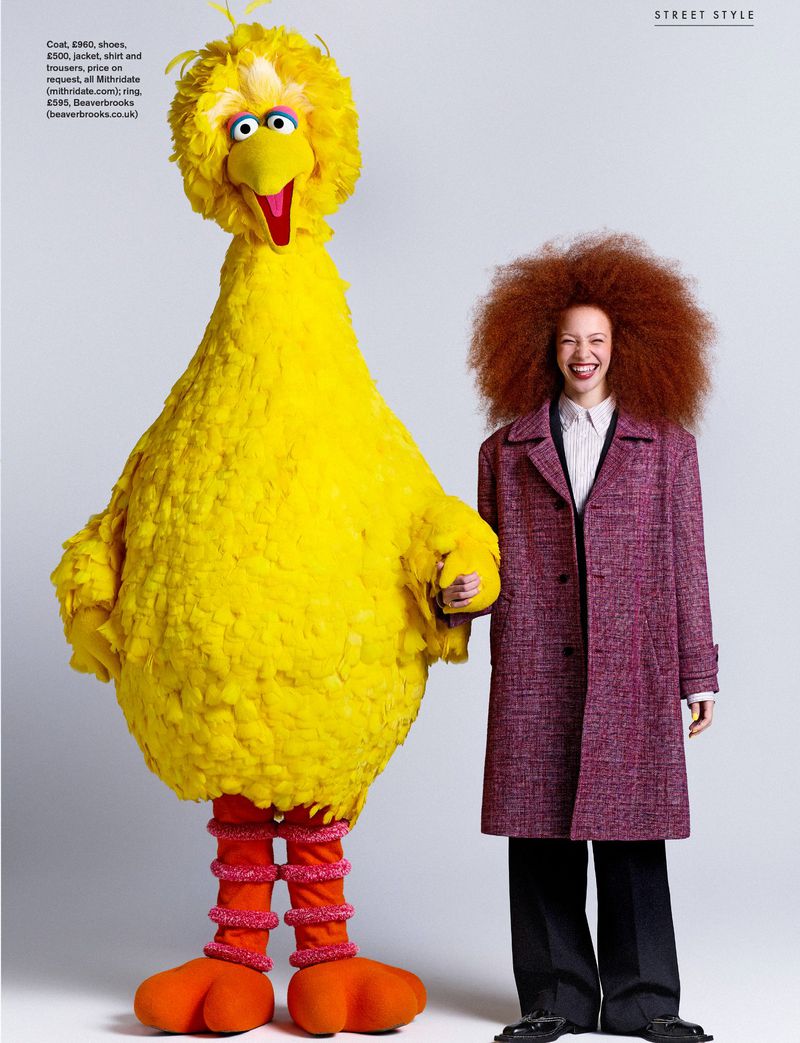 Model Tianna St. Louis in a purple coat holding hands with Sesame Street's Big Bird against a white studio background.