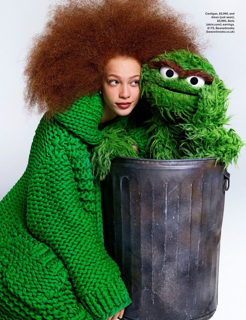 Model Tianna St. Louis in a chunky green knit cardigan posing next to Oscar the Grouch in a trash can against a white backdrop.