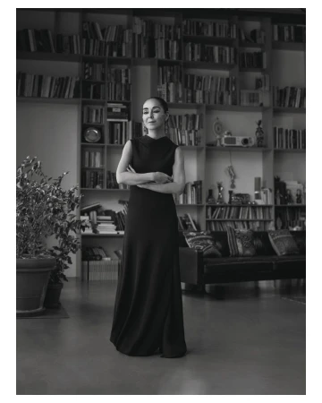 Black and white portrait of artist Shirin Neshat standing in a sleeveless black gown in front of a large bookshelf.