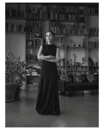 Black and white portrait of artist Shirin Neshat standing in a sleeveless black gown in front of a large bookshelf.