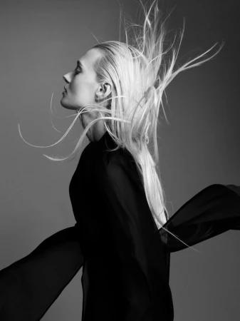 Black and white profile portrait of a woman in a flowing black garment with windswept hair against a plain studio background.