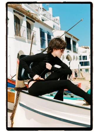 Model Estrella Gomez wearing an all-black long-sleeved outfit and sunglasses while perched on a white boat in a seaside town.