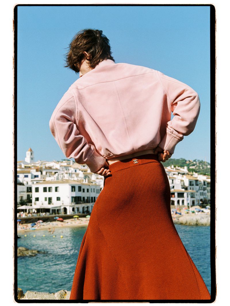 Model Estrella Gomez seen from behind wearing a light pink jacket and rust orange knit skirt overlooking a coastal town.