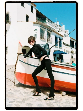Model Estrella Gomez posing in a black long-sleeve cutout outfit and heels against a boat in a sunlit coastal town.