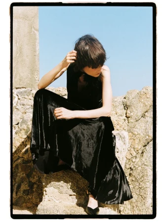 Model Estrella Gomez seated on sunlit stone rocks wearing a sleeveless black velvet maxi dress against a clear blue sky.