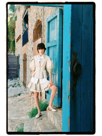 Model Estrella Gomez wearing a cream puff-sleeve peplum jacket and tiered skirt by a rustic stone building with a bright blue door.