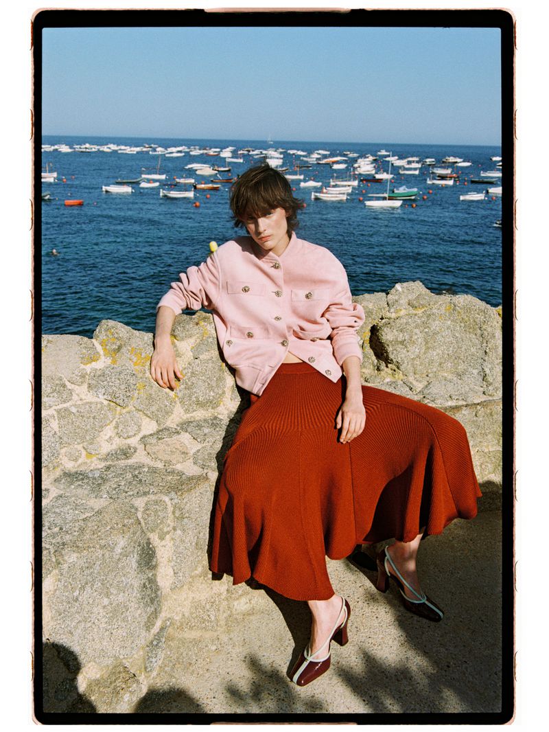 Model Estrella Gomez sitting on a stone wall overlooking the sea wearing a pale pink jacket and burnt orange midi skirt.