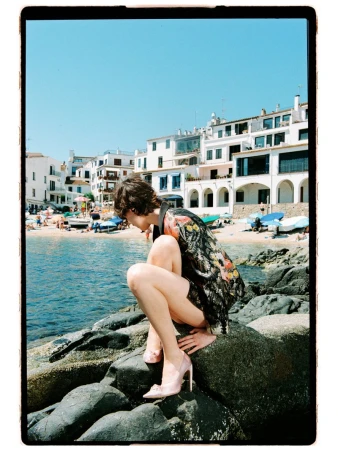 Model Estrella Gomez crouching on coastal rocks wearing a patterned dark blazer and light pink pointed stilettos by the sea.