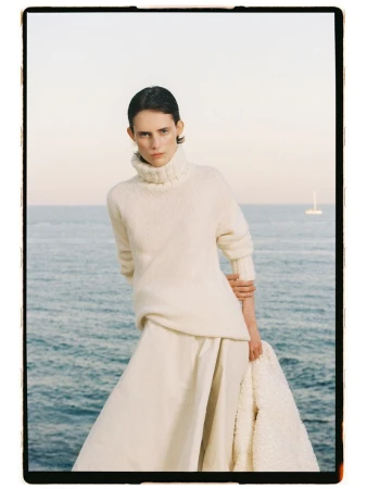 Model Estrella Gomez in an ivory turtleneck sweater and skirt standing against an ocean horizon.