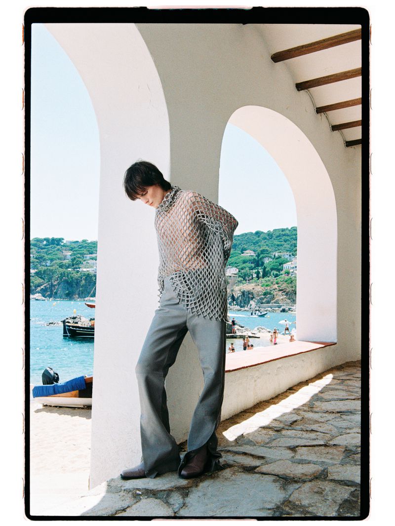 Model posing under white arches wearing an open silver mesh knit top and grey flared trousers overlooking a coastal beach.