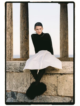 Model Estrella Gomez wearing a black turtleneck, white flared skirt, and fluffy black shoes while seated between stone columns.