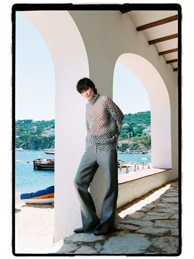 Model Estrella Gomez wearing a grey mesh turtleneck and wide-leg trousers under white arches overlooking a coastal bay.