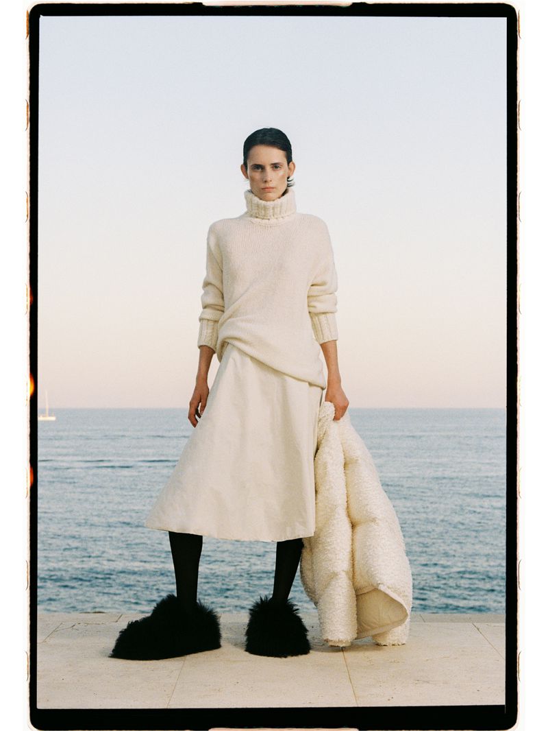 Model Estrella Gomez wearing a cream turtleneck sweater, midi skirt, and black fuzzy slippers standing by the ocean.