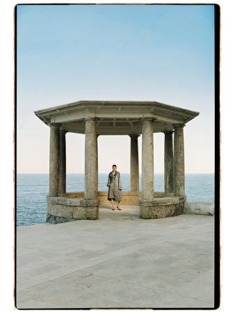 Model Estrella Gomez posing inside an open stone pavilion overlooking the sea in a grey coat for Marie Claire Czech Republic.