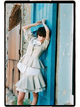 Estrella Gomez leaning against a vivid blue wooden door wearing a beige peplum jacket and pleated skirt.