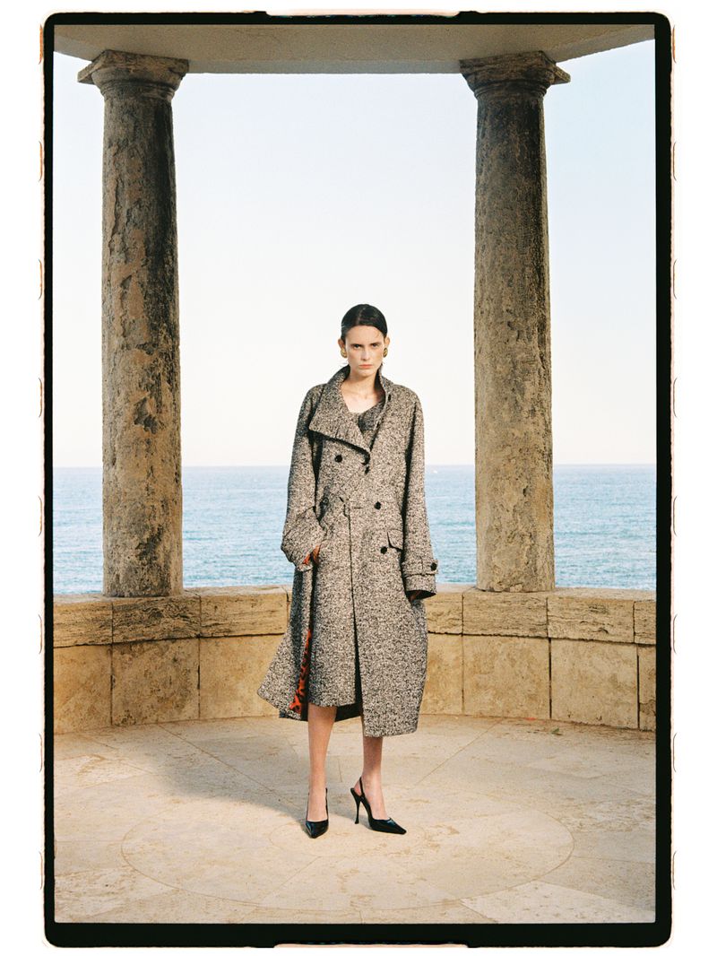 Model in a grey tweed trench coat and black slingback heels standing between stone columns overlooking the ocean.