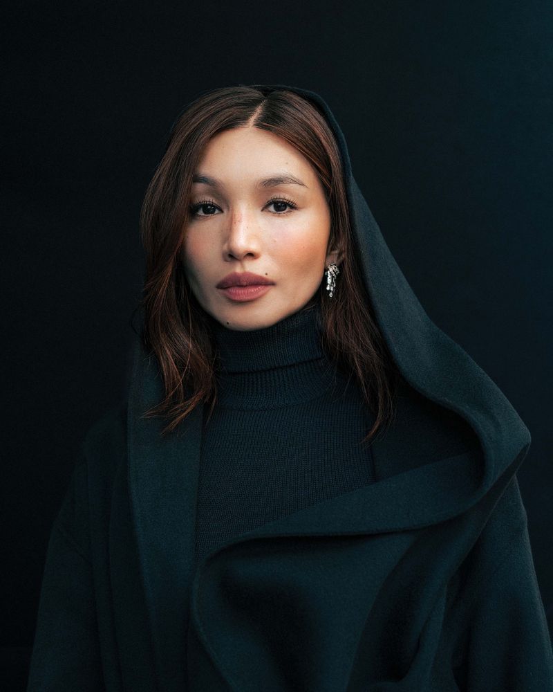Gemma Chan wearing a dark draped hooded coat and turtleneck against a black studio backdrop.