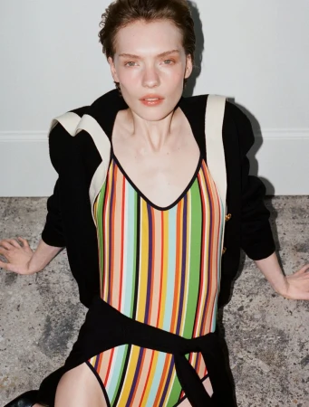 Model Penelope Ternes seated on stone flooring in a vertical striped swimsuit and black cardigan against a white wall.