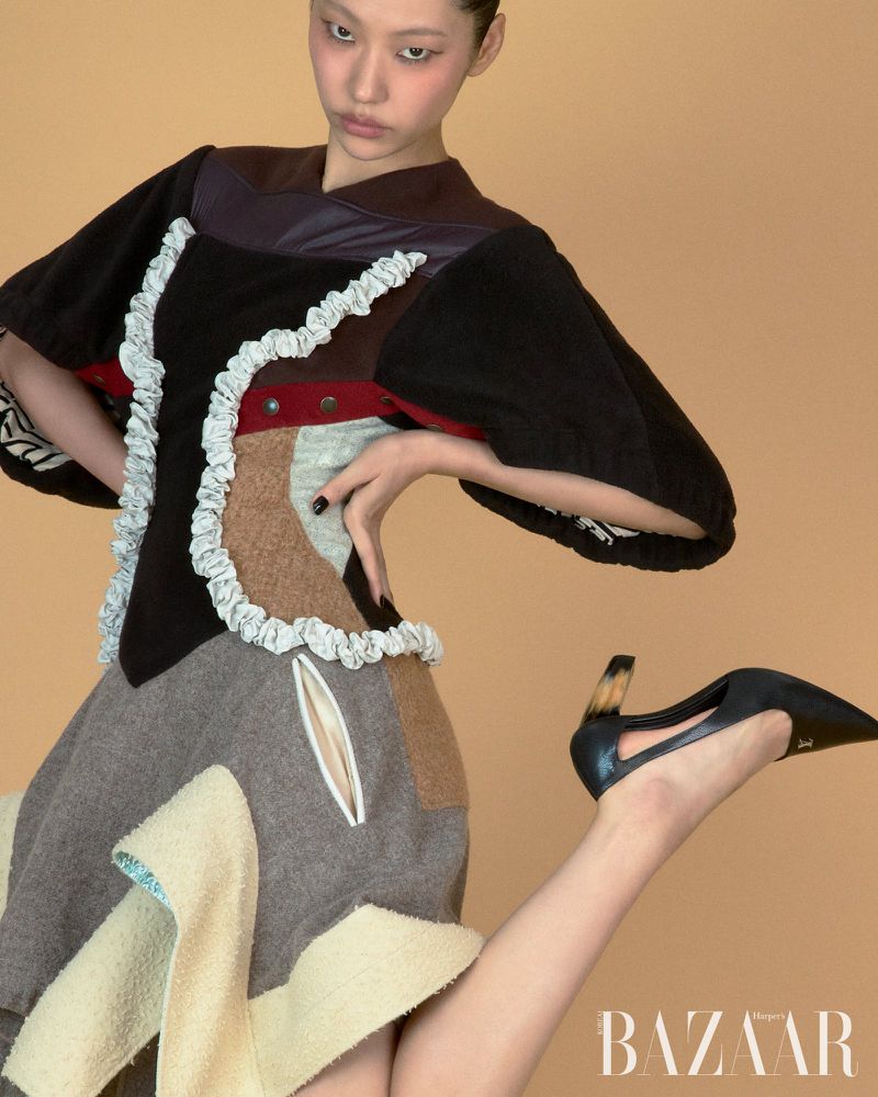 Model Heejung Park posing in a structural patchwork dress and black sculpted heels against a warm beige seamless studio backdrop.