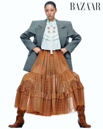Model Heejung Park posing in an oversized grey blazer, white ruffled blouse, orange plaid maxi skirt, and brown boots on white backdrop.