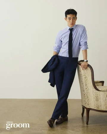Man in a light blue striped shirt, navy necktie, and trousers holding a blazer while leaning against a vintage armchair.