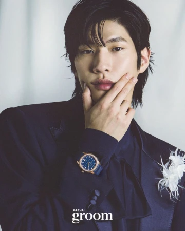 East Asian man in a dark navy suit and gold luxury watch with hand on chin against a white draped backdrop.