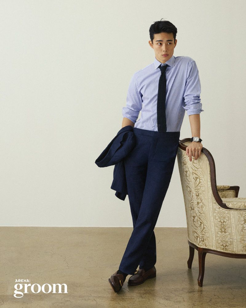 Model in a light blue dress shirt and navy trousers leaning on an upholstered armchair against an off-white studio background.