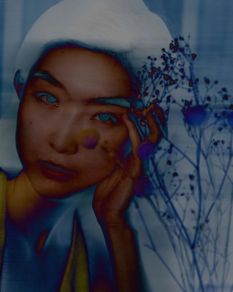 Editorial close-up portrait of an East Asian woman resting her hand on her cheek beside delicate dried branches with a cool blue effect.