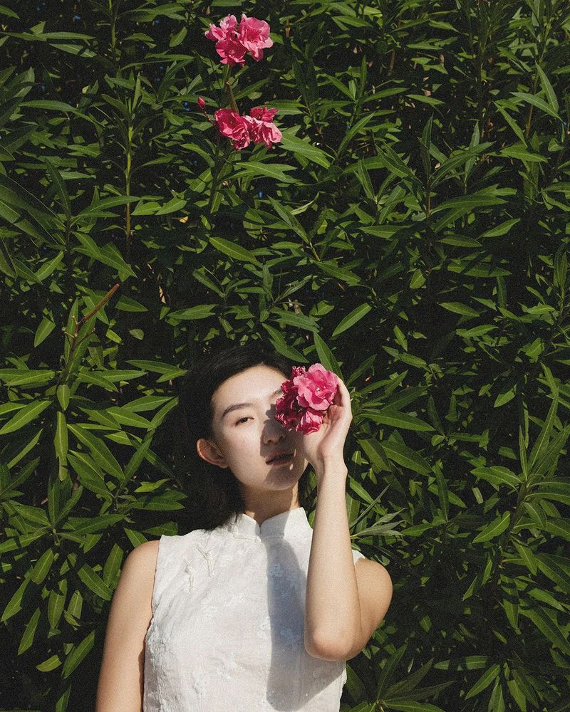 An East Asian woman in a sleeveless white dress holds pink flowers over her eye in front of green foliage.