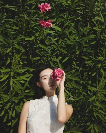 An East Asian woman in a sleeveless white dress holds pink flowers over her eye in front of green foliage.