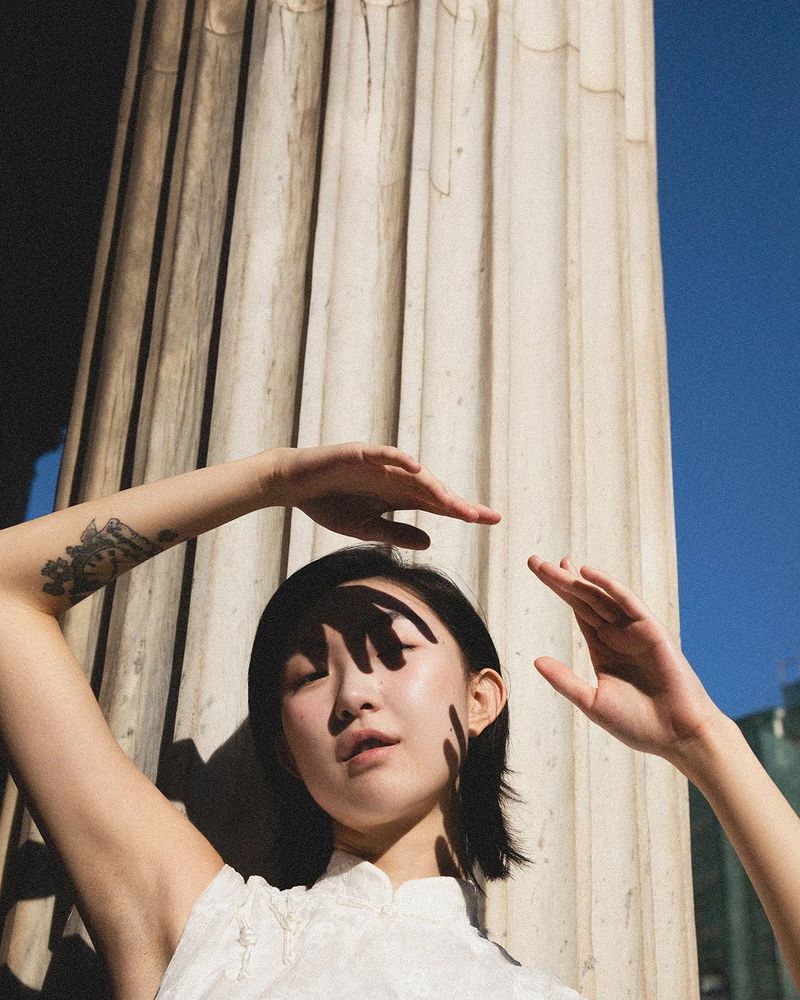 East Asian woman posing with raised arms casting hand shadows across her face against a fluted marble column in bright sunlight.