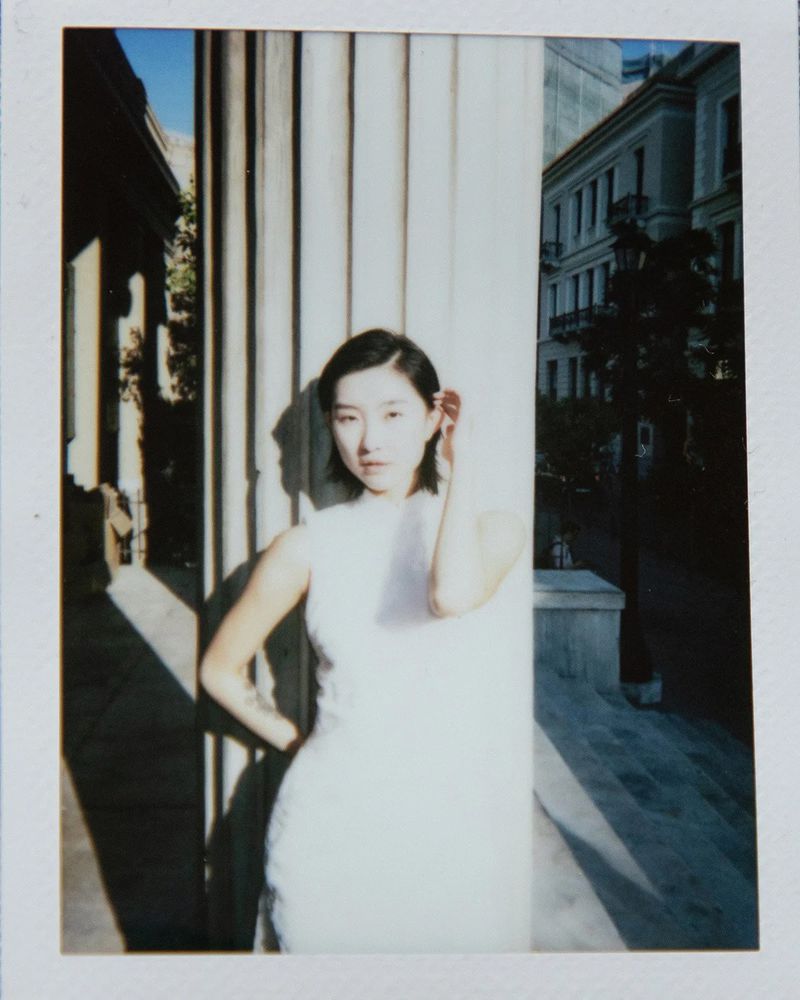Polaroid portrait of an East Asian woman wearing a white sleeveless dress and posing against a fluted classical pillar in bright sunlight.