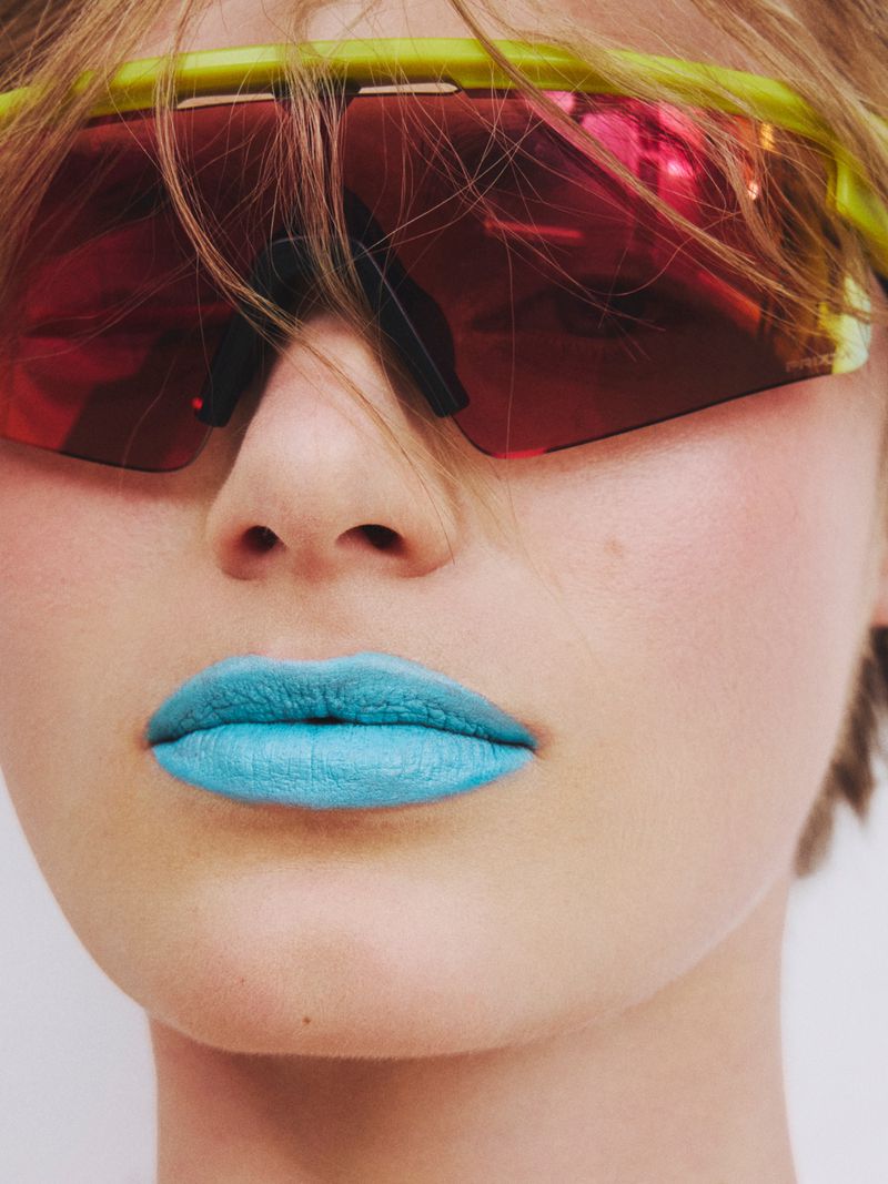 Close-up portrait of a model wearing bright blue lipstick and neon yellow sports sunglasses with red mirrored lenses.