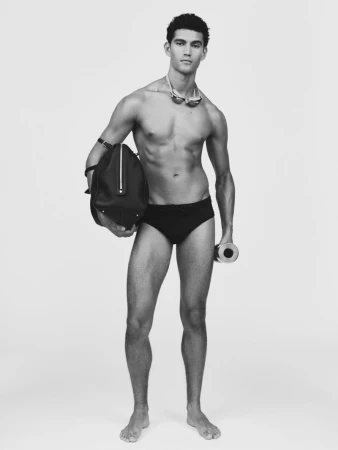Model Bodhi Heeck posing in black swim briefs with a duffle bag and goggles against a seamless studio background.