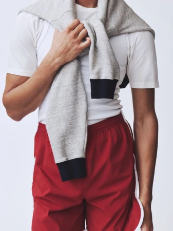 Cropped torso of a model wearing red running shorts with white trim and a grey sweatshirt draped over shoulders against a white background.