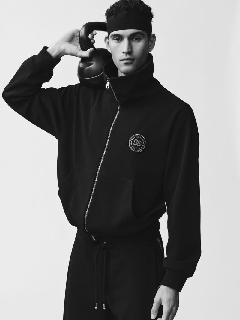 Model Bodhi Heeck wearing a black zip-up jacket and headband while holding a kettlebell against a white background.