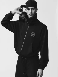 Model Bodhi Heeck wearing a black zip-up jacket and headband while holding a kettlebell against a white background.