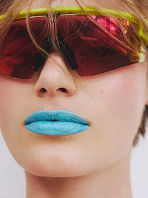 Close-up portrait of a model wearing bright turquoise lipstick and neon lime shield sunglasses with red mirrored lenses.