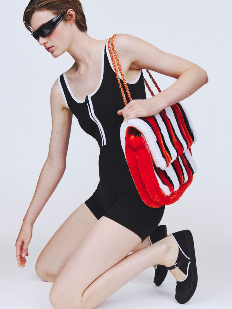 Model kneeling in a black athletic romper with white trim, dark wraparound sunglasses, and an oversized striped plush shoulder bag.