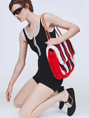 Model kneeling in a black athletic romper with white trim, dark wraparound sunglasses, and an oversized striped plush shoulder bag.