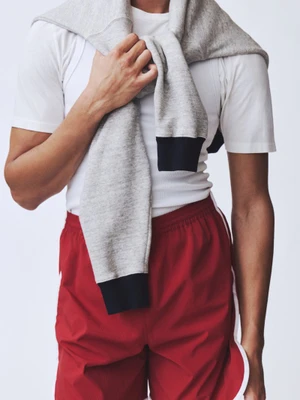Model wearing red athletic shorts with white trim, a white t-shirt, and a grey sweatshirt draped over shoulders against a white backdrop.