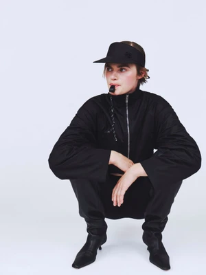Model Bodhi Heeck wearing an all-black sporty jacket, visor, and boots while crouching with a whistle in mouth against a white backdrop.