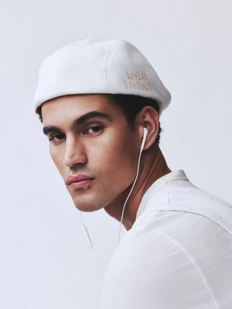 Model Bodhi Heeck wearing a white embroidered cap and wired earphones against a pale studio backdrop.