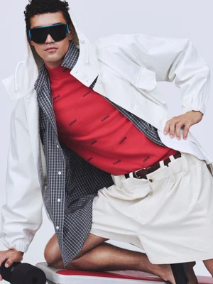 Model wearing black shield sunglasses, a red mock-neck top, a white hooded jacket, and white shorts against a light background.