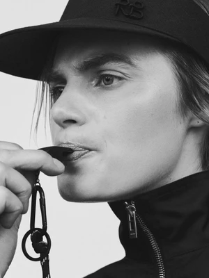 Close-up black and white portrait of a model blowing a whistle while wearing a black cap and zip-up jacket.