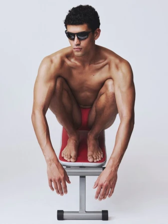 Model Bodhi Heeck crouching barefoot on a red gym bench wearing sunglasses and red briefs against a white studio background.